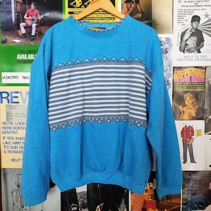 May include: A blue sweatshirt with a white and gray striped pattern and a geometric design. The sweatshirt has long sleeves and a crew neck.