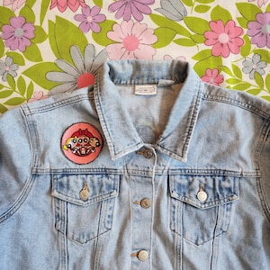 May include: A light blue denim jacket with a pink and red embroidered patch of the Powerpuff Girls cartoon characters. The patch is on the left side of the jacket, above the pocket.