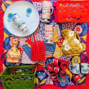 May include: A collection of vintage kitchenware and decorative items, including a floral-decorated plate, juice mugs, a pitcher, napkin rings, and a kangaroo figurine. The items are arranged on a colorful patchwork quilt.