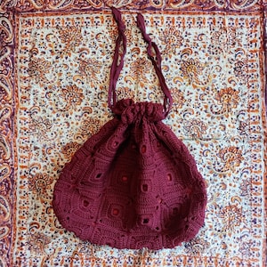 May include: A maroon crocheted drawstring bag with a square pattern and a long strap. The bag is sitting on a patterned fabric.