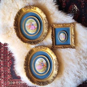 May include: Three ornate gold-colored picture frames with blue velvet inserts. Each frame holds a small oval or rectangular porcelain plaque depicting a couple in a garden setting. The frames are arranged on a white fluffy surface.