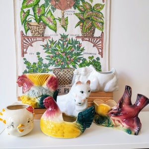 May include: A collection of five vintage ceramic planters, each featuring a different design. The planters are in various colors, including white, red, yellow, and green. The planters are arranged on a wooden surface. The planters are decorated with various designs, including a rooster, a cat, a bird, and a flower.