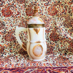 May include: A white alabaster pitcher with a gold metal band around the top. The pitcher has a unique, swirling pattern and a handle. It is sitting on a patterned fabric background.