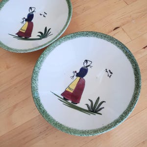 May include: Two white ceramic bowls with green trim and a painted design of a woman in a yellow and red dress holding flowers. The bowls are on a wooden surface.