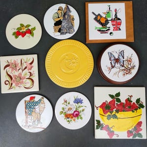 May include: A collection of decorative ceramic coasters and tiles. Designs include strawberries, floral patterns, a butterfly, a yellow plate with a sun and moon, and a person weaving. The coasters and tiles are in various shapes and sizes.
