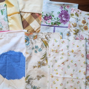 May include: A collection of vintage pillowcases in various patterns and colors. Designs include floral prints with pink, purple, and blue flowers, a plaid pattern, and a solid yellow case. The pillowcases are made of cotton or a similar fabric.