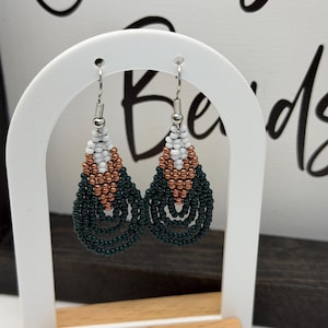 May include: A pair of beaded earrings in a teardrop shape. The earrings feature a pattern of white, copper, and dark green beads. The earrings are displayed on a white arch-shaped stand with a wooden base.