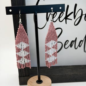 May include: A pair of beaded earrings in a teardrop shape. The earrings are primarily dusty rose with a white geometric pattern. The earrings are displayed on a black stand with a wooden base, against a white background.