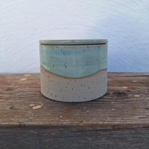 May include: A handmade ceramic jar with a lid. The jar has a cylindrical shape with a light blue top and a speckled beige bottom. The lid matches the top. The jar is sitting on a wooden surface.