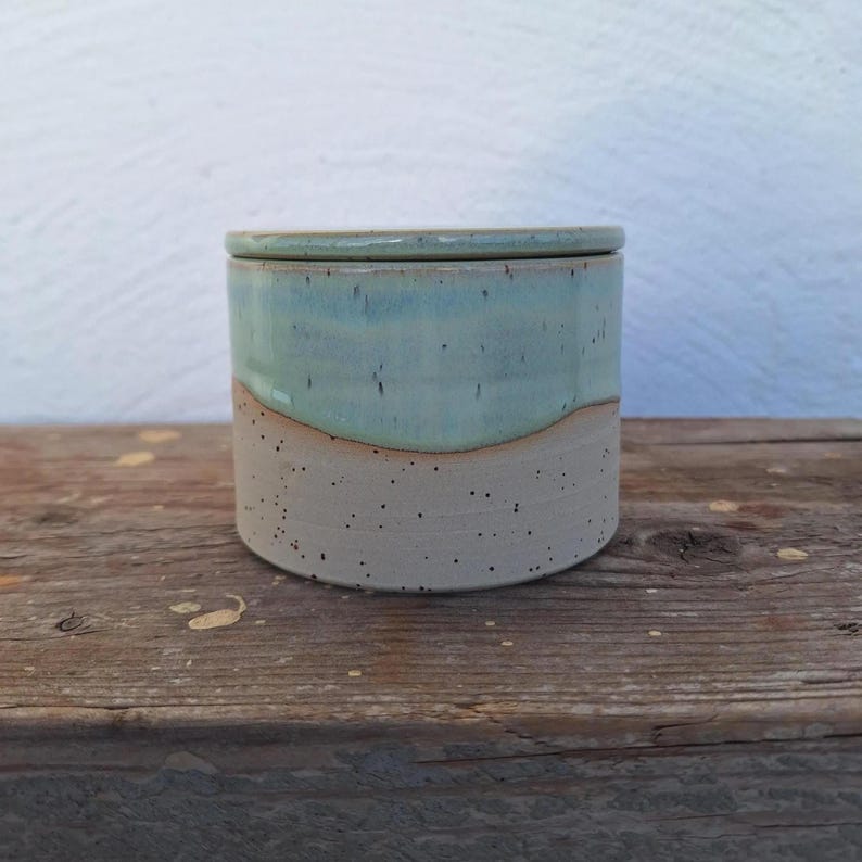 May include: A handmade ceramic jar with a lid. The jar has a cylindrical shape with a light blue top and a speckled beige bottom. The lid matches the top. The jar is sitting on a wooden surface.