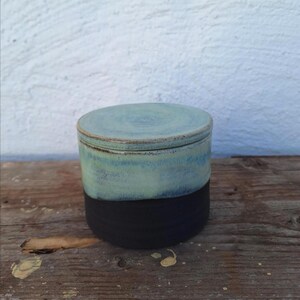 May include: A handmade ceramic jar with a lid. The jar is cylindrical, with a black base and a top section that fades from light blue to a darker teal. The lid is a matching light blue. The jar sits on a wooden surface.