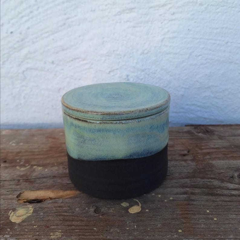 May include: A handmade ceramic jar with a lid. The jar is cylindrical, with a black base and a top section that fades from light blue to a darker teal. The lid is a matching light blue. The jar sits on a wooden surface.