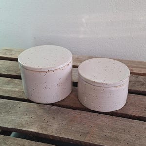 May include: Two ceramic jars with lids. The jars are cylindrical with a speckled, off-white finish and dark brown speckles. They are sitting on a weathered wooden surface.