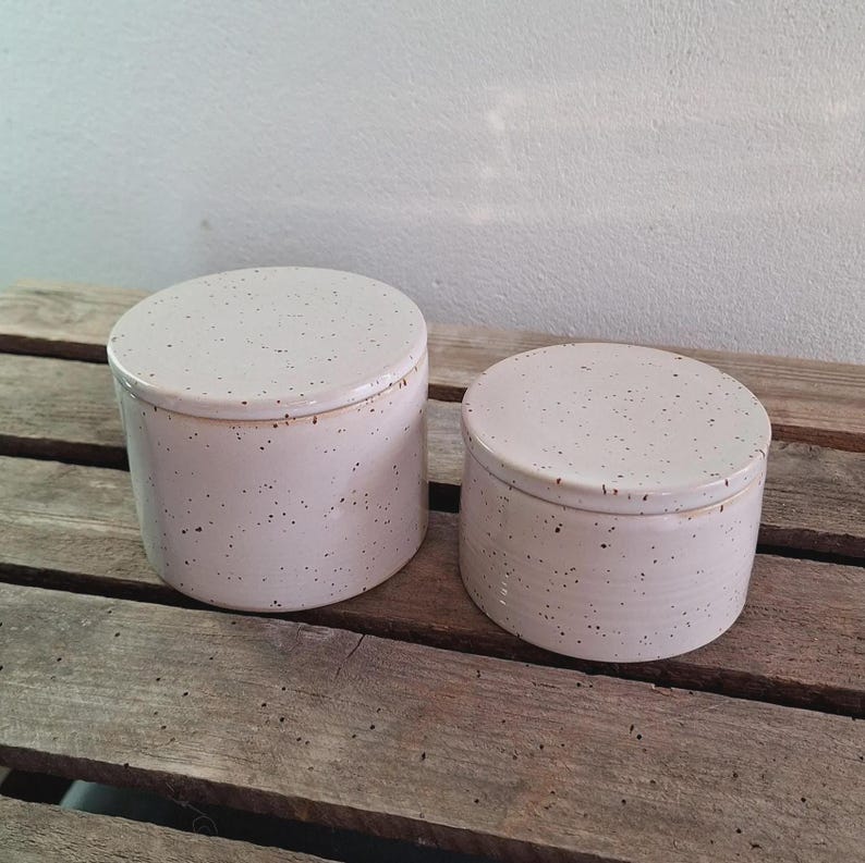 May include: Two ceramic jars with lids. The jars are cylindrical with a speckled, off-white finish and dark brown speckles. They are sitting on a weathered wooden surface.