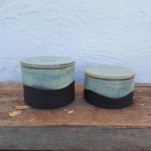 May include: Two ceramic lidded jars. The jars have a two-tone design, with a black base and a light blue top. The lids are also light blue. The jars are sitting on a wooden surface.