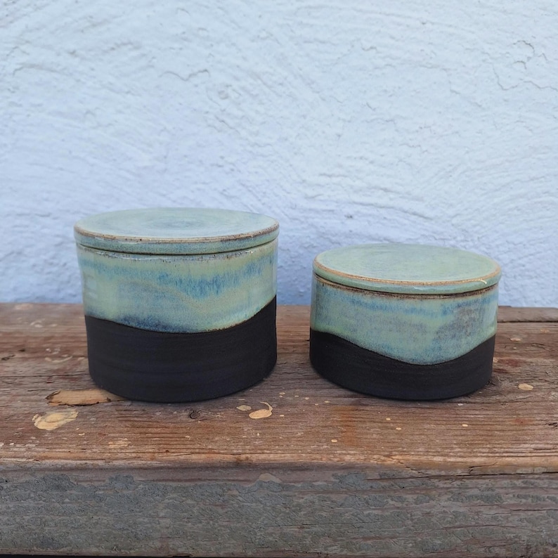 May include: Two ceramic lidded jars. The jars have a two-tone design, with a black base and a light blue top. The lids are also light blue. The jars are sitting on a wooden surface.