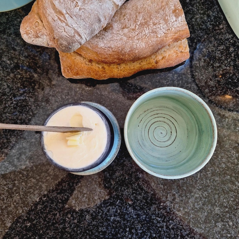 May include: A butter dish with a wooden knife, filled with butter, next to an empty ceramic bowl. A loaf of bread is in the background. The butter dish has a blue rim and the bowl is light green with a spiral design.