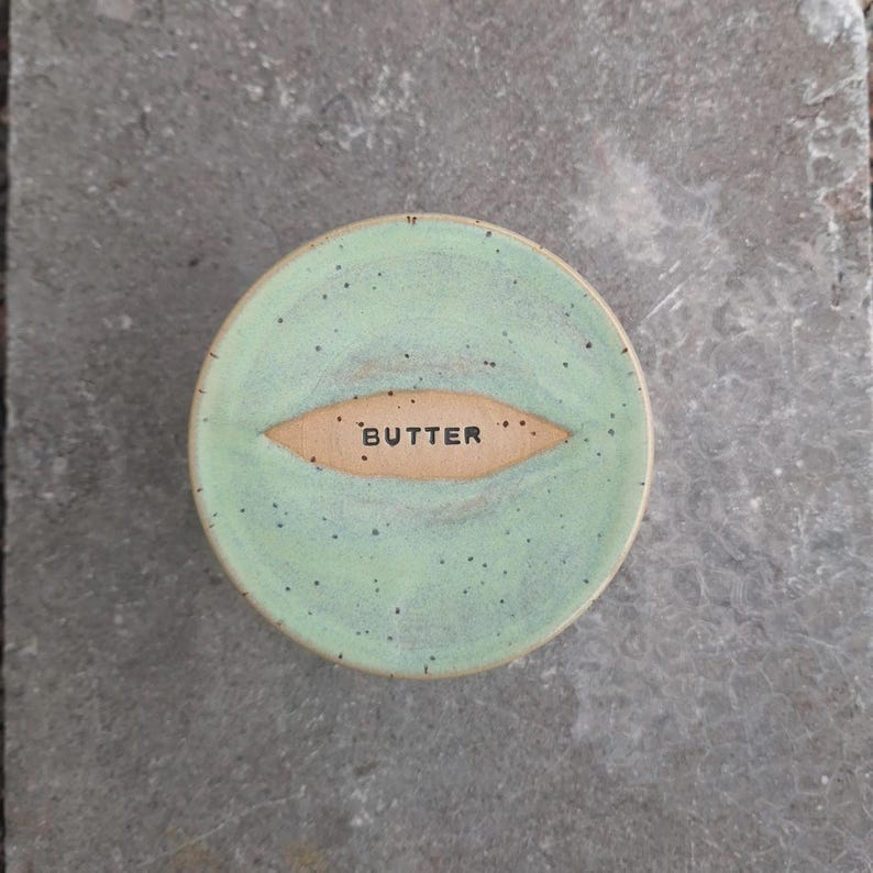 May include: A round, ceramic butter dish with a light green glaze and speckled finish. The word "BUTTER" is inscribed in a tan oval in the center. The dish is set on a gray stone surface.