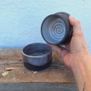 May include: Two handmade ceramic bowls. One bowl is dark blue with a black base, the other is held up, revealing a spiral design. The bowls are on a wooden surface against a light blue wall.