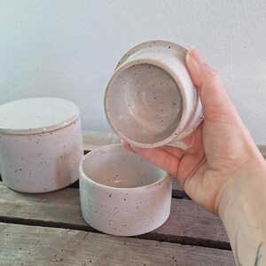 May include: A set of three speckled ceramic containers. One container has a lid, another is open, and the third is being held. The containers are off-white with small, dark speckles. The containers are cylindrical in shape.