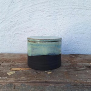 May include: A handmade ceramic jar with a lid. The jar is cylindrical, with a black base and a top section that fades from light blue to a darker blue-green. The lid matches the top section. The jar sits on a wooden surface.