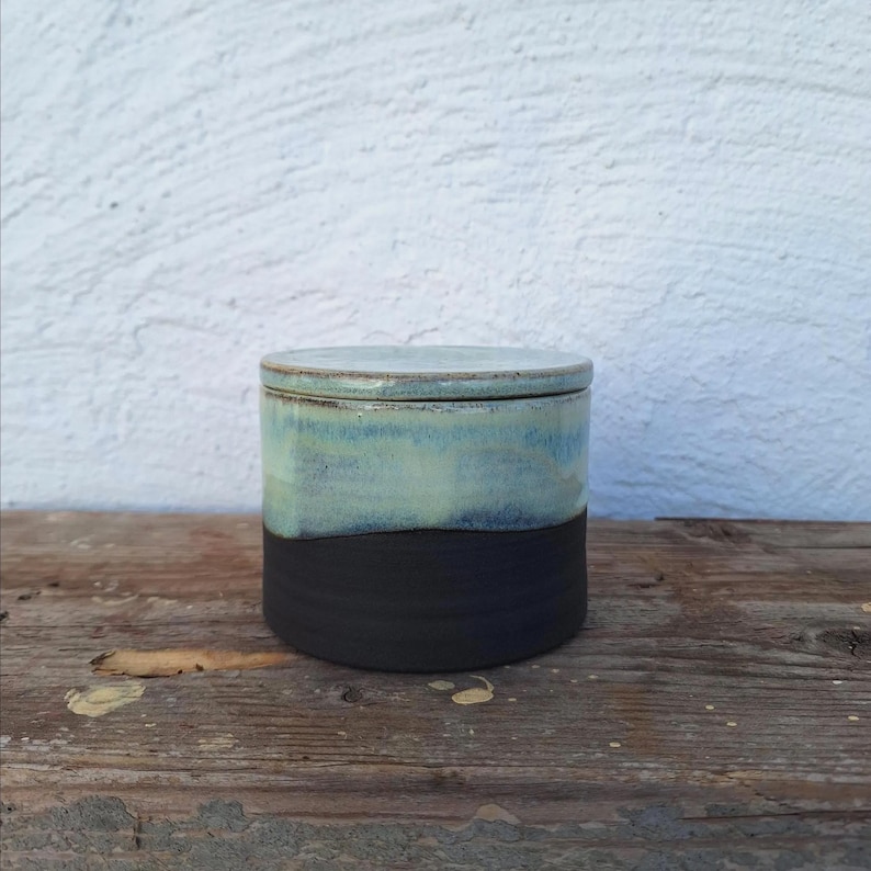 May include: A handmade ceramic jar with a lid. The jar is cylindrical, with a black base and a top section that fades from light blue to a darker blue-green. The lid matches the top section. The jar sits on a wooden surface.