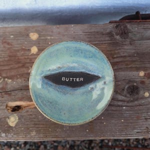 May include: A round, ceramic butter dish with a turquoise glaze. The dish has a dark, oval-shaped indentation in the center with the word "BUTTER" in white lettering. The dish sits on a weathered wooden surface.