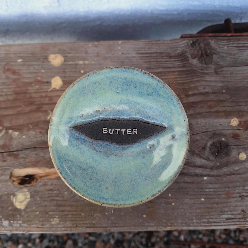 May include: A round, ceramic butter dish with a turquoise glaze. The dish has a dark, oval-shaped indentation in the center with the word "BUTTER" in white lettering. The dish sits on a weathered wooden surface.