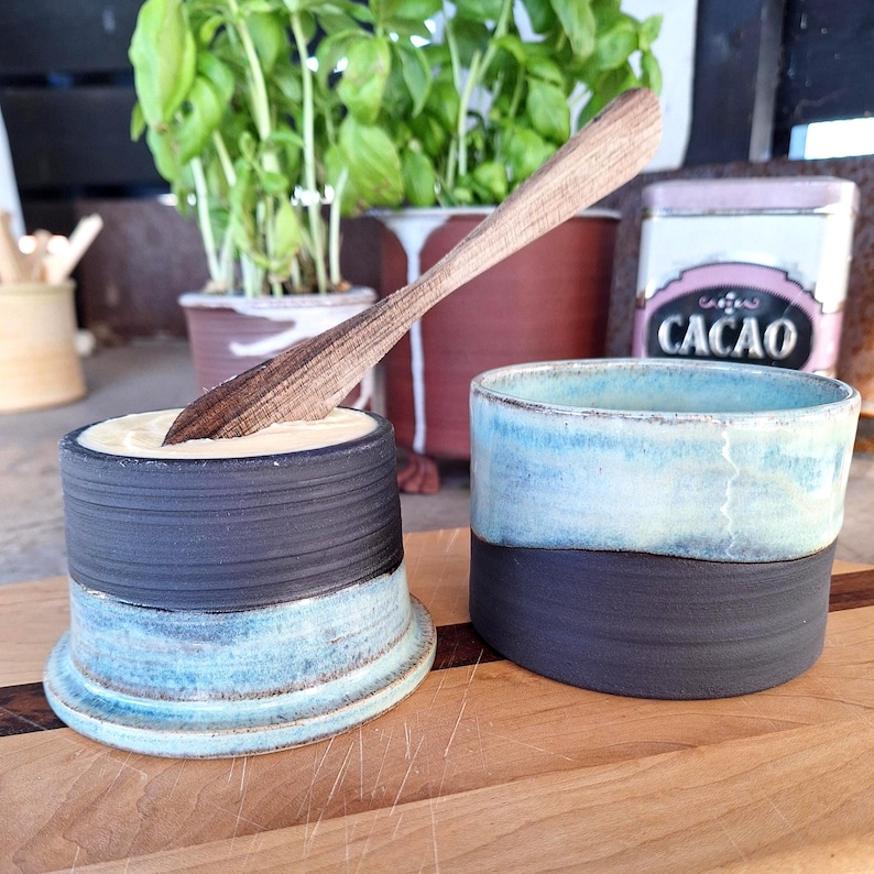 May include: Two handmade ceramic butter keepers with a wooden butter knife. One keeper is filled with butter. The keepers have a two-tone design, with a dark blue base and a light blue top. The wooden knife is resting in the butter.