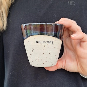 May include: A ceramic cup with a speckled off-white base and a dark, glossy, iridescent rim. The text ON FIRE! is stamped in black capital letters on the front of the cup. The vessel has a tapered shape and a smooth, rounded top edge.