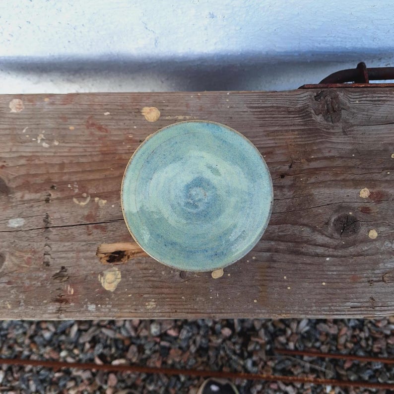 May include: A handmade ceramic plate with a light blue glaze. The plate has a circular shape with a slightly raised edge. The glaze has a swirl pattern in shades of blue and green. The plate is sitting on a weathered wooden surface.
