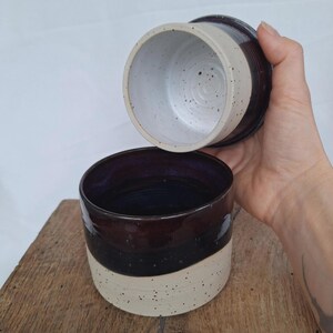 May include: A two-piece ceramic butter dish with a dark brown and cream speckled design. The bottom piece is a bowl shape with a dark blue interior. The lid is a matching dark brown and cream speckled design.