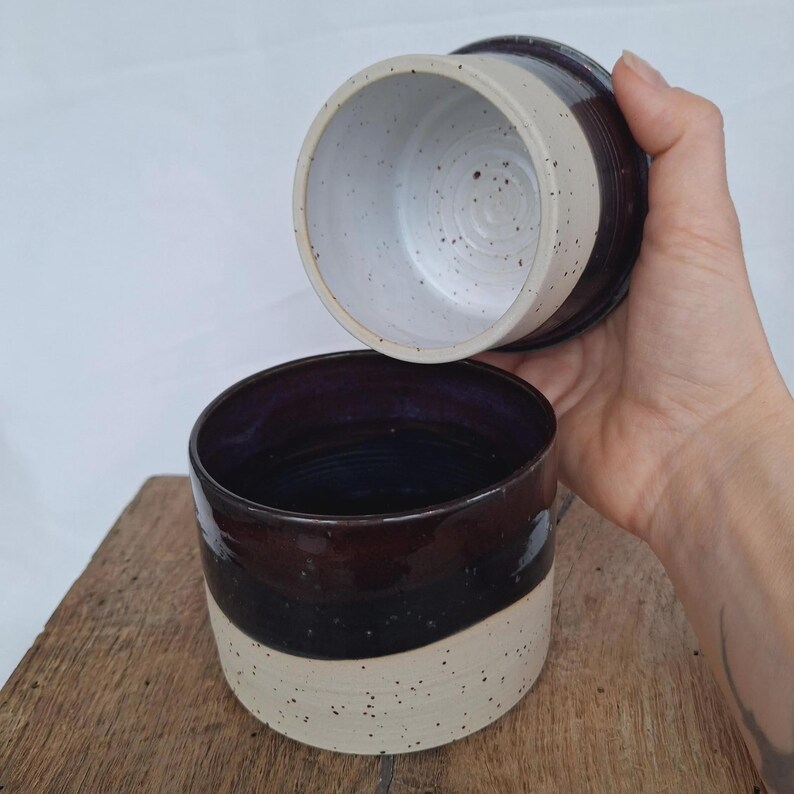 May include: A two-piece ceramic butter dish with a dark brown and cream speckled design. The bottom piece is a bowl shape with a dark blue interior. The lid is a matching dark brown and cream speckled design.