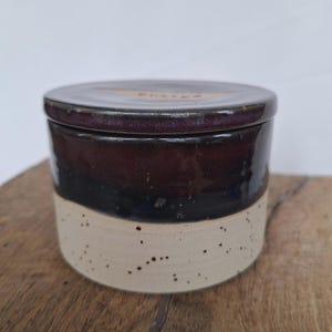May include: A ceramic butter dish with a lid. The dish is cylindrical with a dark brown top, a black middle section, and a speckled beige base. The lid has the word "butter" etched on it.