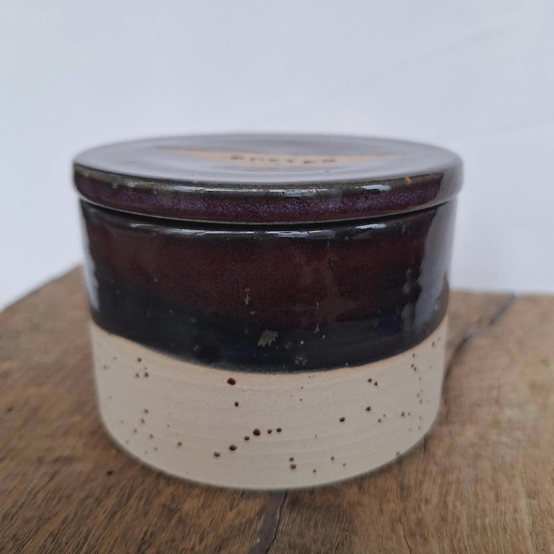 May include: A ceramic butter dish with a lid. The dish is cylindrical with a dark brown top, a black middle section, and a speckled beige base. The lid has the word "butter" etched on it.