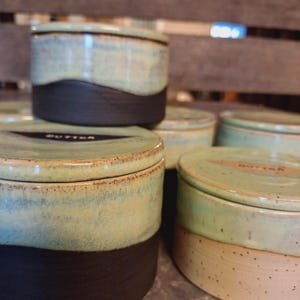 May include: Handmade ceramic butter dishes with lids. The dishes have a two-tone design, with a dark brown base and a light blue-green top. The word "BUTTER" is printed on the lid. The dishes are stacked, showcasing their unique, rustic appearance.
