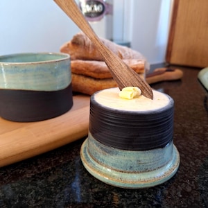 Puede incluir: Una mantequera de cerámica hecha a mano con una taza a juego. La mantequera es cilíndrica con un diseño de dos tonos, con una parte superior azul-negro oscuro y una base azul-verde claro. Un cuchillo de mantequilla de madera descansa en el plato, con una porción de mantequilla.