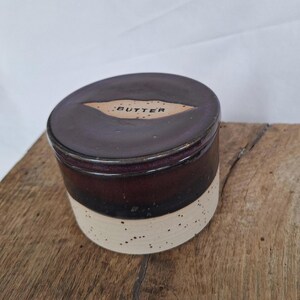 May include: A ceramic butter dish with a dark brown lid and a cream-colored speckled base. The lid has a label that reads "BUTTER". The dish is cylindrical and sits on a wooden surface.