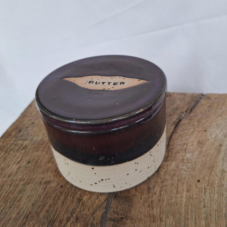 May include: A ceramic butter dish with a dark brown lid and a cream-colored speckled base. The lid has a label that reads "BUTTER". The dish is cylindrical and sits on a wooden surface.
