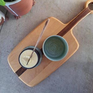 May include: A wooden serving board with a handle, featuring a dark wood stripe. It holds a small, light blue bowl with a spiral design, a small bowl with a creamy substance, and a wooden spoon. A terracotta pot is visible in the background.