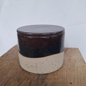 May include: A ceramic jar with a lid. The jar is cylindrical with a dark brown top, a black middle section, and a speckled beige bottom. The lid is a matching dark brown color. The jar is sitting on a wooden surface.