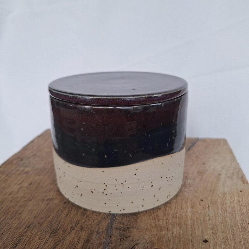 May include: A ceramic jar with a lid. The jar is cylindrical with a dark brown top, a black middle section, and a speckled beige bottom. The lid is a matching dark brown color. The jar is sitting on a wooden surface.