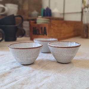 Op de afbeelding: Drie kleine keramische kommen met een gespikkeld wit glazuur en een opvallende bruine rand. De kommen hebben een conische vorm met een gladde binnenkant en een licht getextureerd buitenoppervlak. Elke kom heeft een consistent gespikkeld patroon.