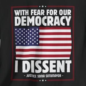 Può includere: T-shirt nero con la bandiera americana e la scritta "WITH FEAR FOR OUR DEMOCRACY I DISSENT - JUSTICE SONIA SOTOMAYOR." Il design include colori rosso, bianco e blu.