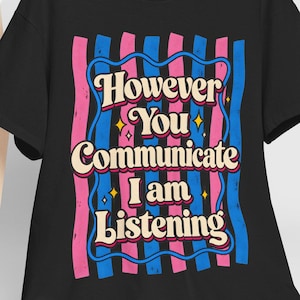 Può includere: Una t-shirt nera a maniche corte con una grafica a strisce verticali rosa e blu. Il testo "However You Communicate I am Listening" è scritto in color crema, circondato da un bordo blu ondulato e da piccole stelle gialle decorative.