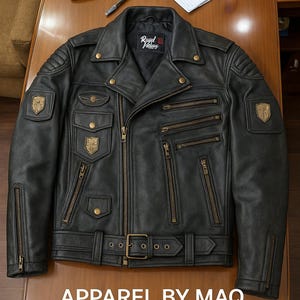 Pode incluir: Jaqueta de motociclista de couro preto com vários bolsos com zíper, cinto e patches decorativos. A jaqueta tem um estilo motociclista clássico com gola entalhada e ombreiras. O texto "Royal Falcons" é visível na gola. O texto "APPAREL BY MAQ" é visível na parte inferior.