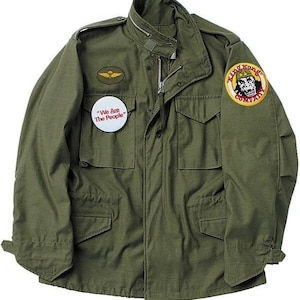 May include: Olive green military-style jacket with a zipper closure, multiple pockets, and a stand-up collar. Features a small patch with a gold emblem and a circular pin that reads "We Are The People." A "King Kong Company" patch is on the right sleeve.