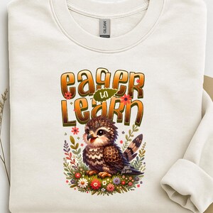 May include: Cream-colored sweatshirt featuring a colorful illustration of an owl surrounded by flowers. The text "Eager to Learn" is printed above the owl in a stylized font. The sweatshirt has a crew neck and long sleeves.