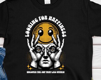 May include: Black t-shirt with a graphic design featuring a yellow smiley face with eyes and hands, and the text "LOOKING FOR HAPPINESS". The shirt is paired with blue jeans on a white wooden surface.