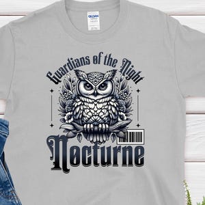 May include: A light gray t-shirt featuring a detailed black and white owl design with the text "Guardians of the Night" and "Nocturne". The shirt is paired with blue jeans and a sprig of greenery.